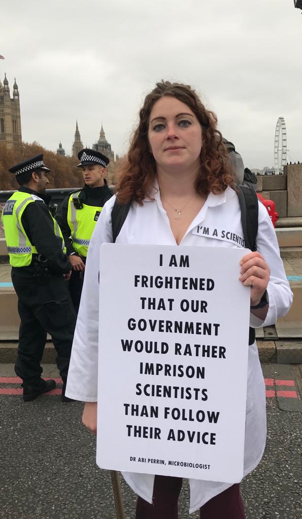Dr Abi Perrin wearing a lab coat holding a placard saying "I AM FRIGHTENED THAT OUR GOVERNMENT WOULD RATHER IMPRISON SCIENTISTS THAN FOLLOW THEIR ADVICE."